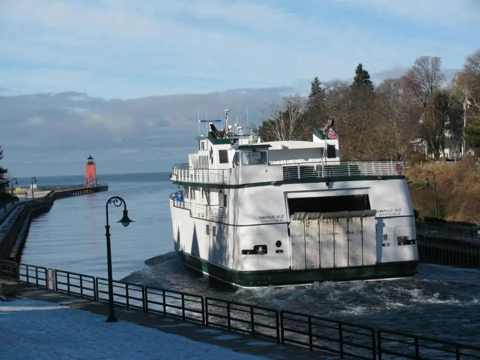 A white-and-green ferry passes between snow-covered piers, with a red lighthouse beyond.