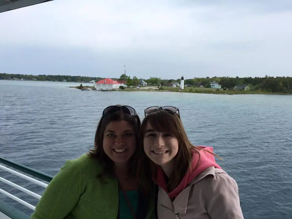 Two women smile on a boat deck, with a lighthouse, houses, and wooded shoreline behind them.