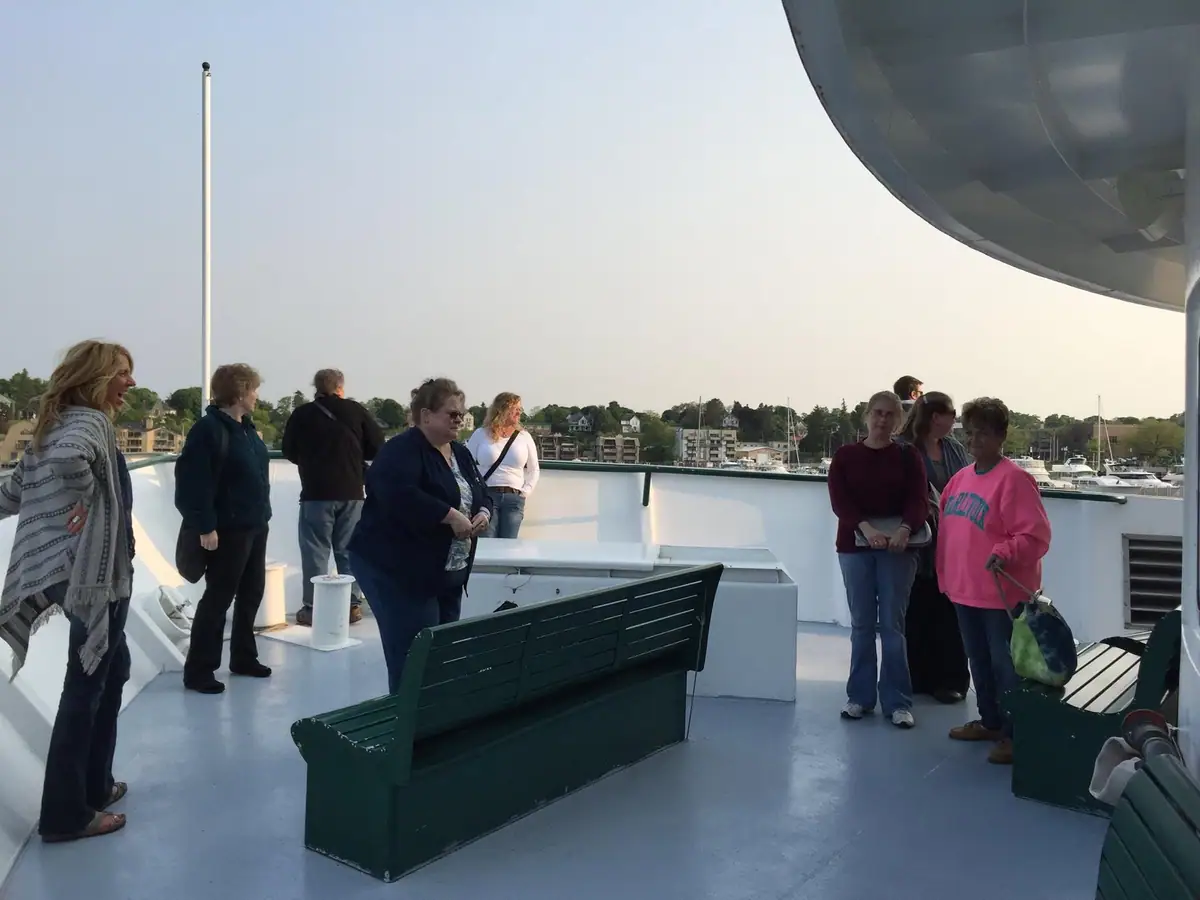 Passengers gather on an open ferry deck overlooking a harbor and wooded shoreline.