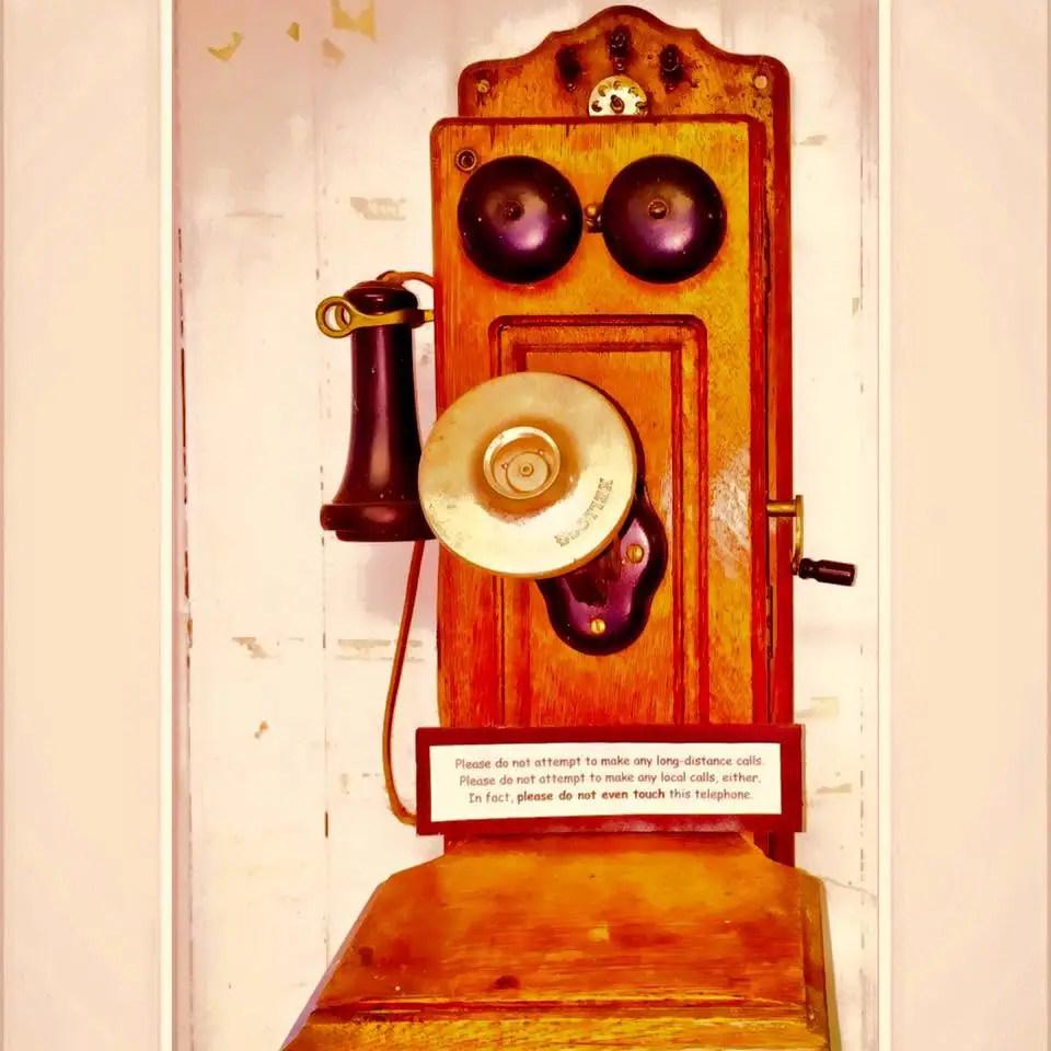 A wall-mounted wooden telephone with twin bells, a side handset, crank, and warning sign.