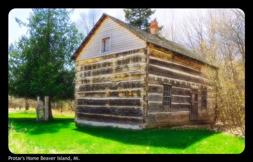 A log house with a gabled roof, brick chimney, and small wooden gate beside a grassy yard.