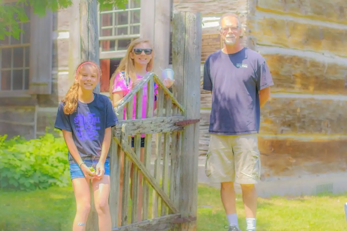 Three people pose beside a weathered wooden gate in front of a log building.