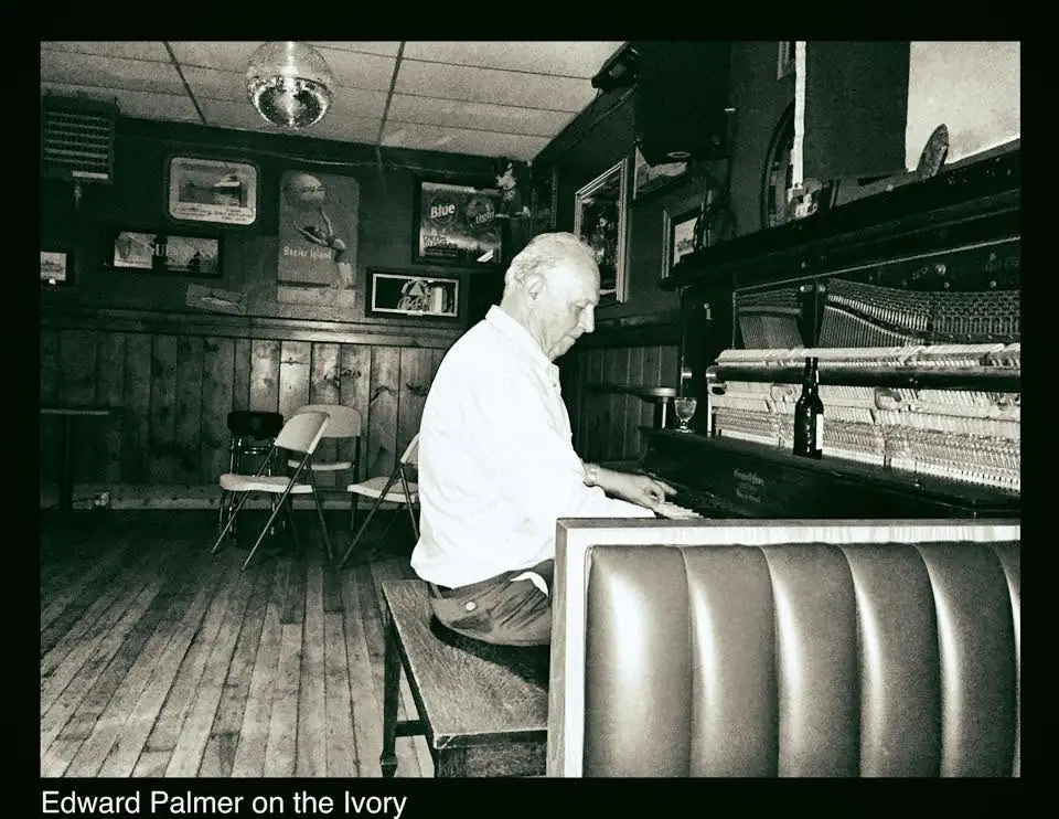 Black-and-white photograph of a man playing an upright piano in a wood-paneled bar or lounge.