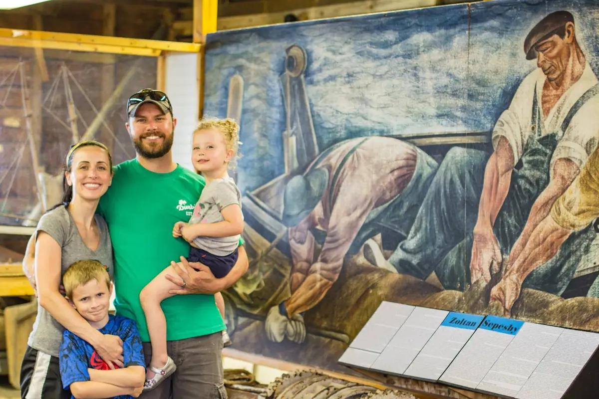 Four people pose beside a large mural of workers aboard a boat inside an exhibit space.