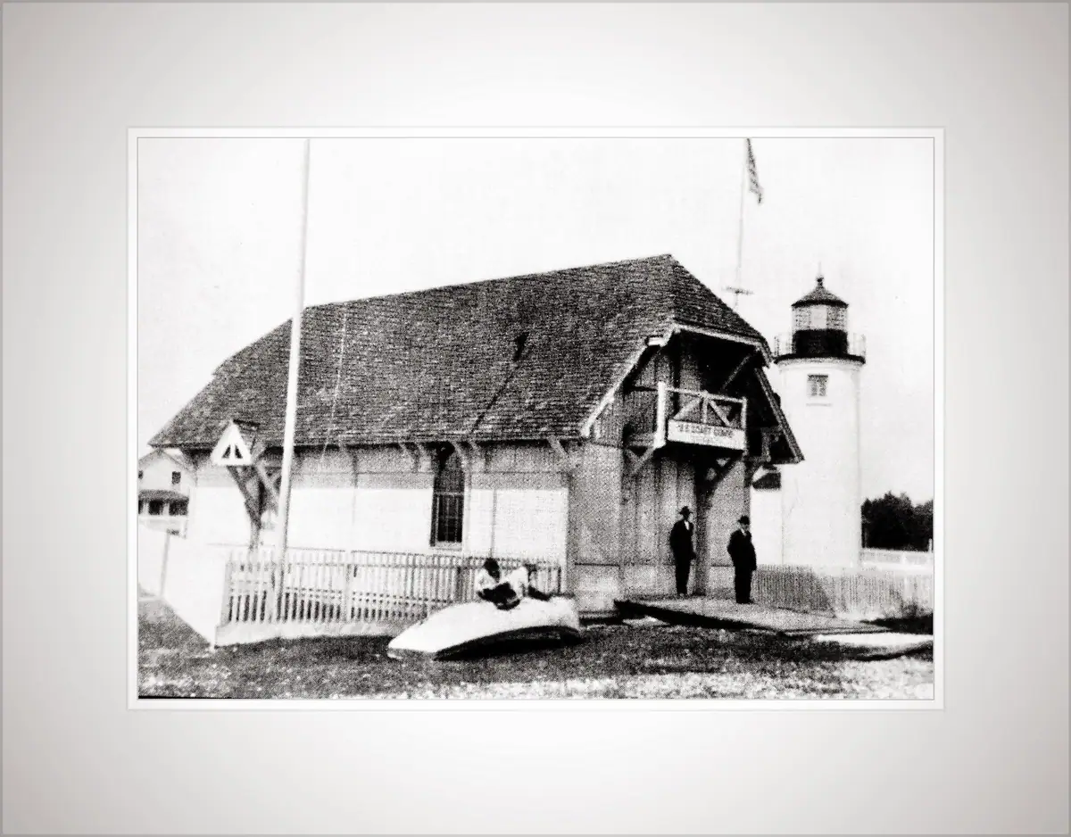 Black-and-white photograph of a shingled station beside a lighthouse, with three people and an overturned boat.