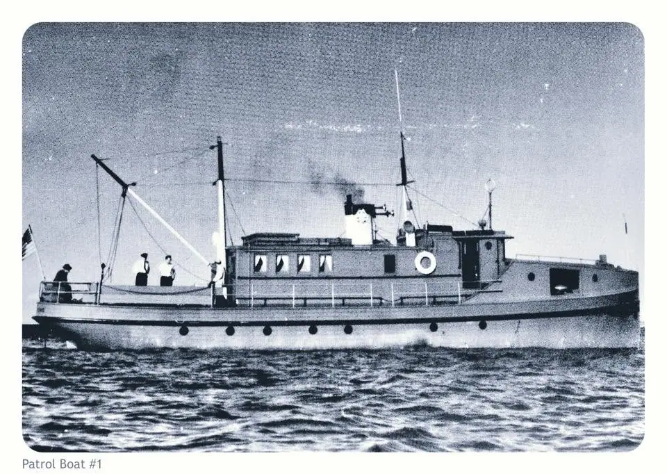 Black-and-white side view of a patrol boat underway, with several people aboard and a flag at the stern.