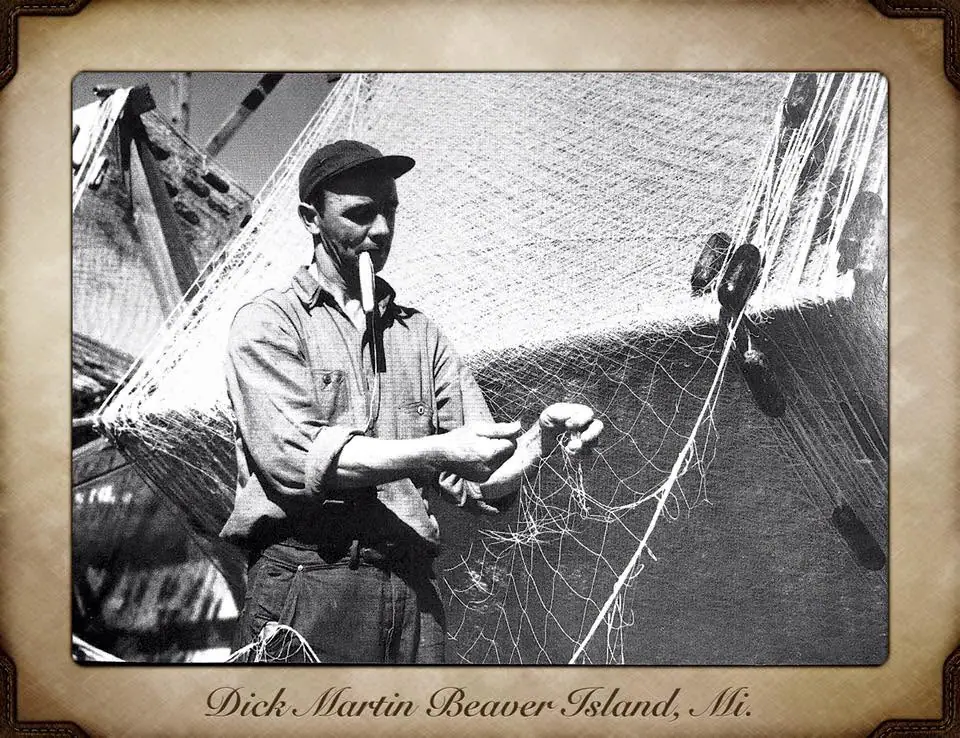 A man repairs a fishing net while holding a twine needle in his mouth.
