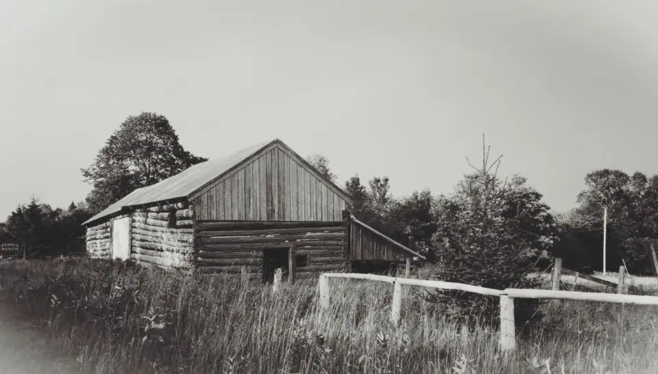 Black-and-white photograph of a log building beside a grassy field and wooden fence.