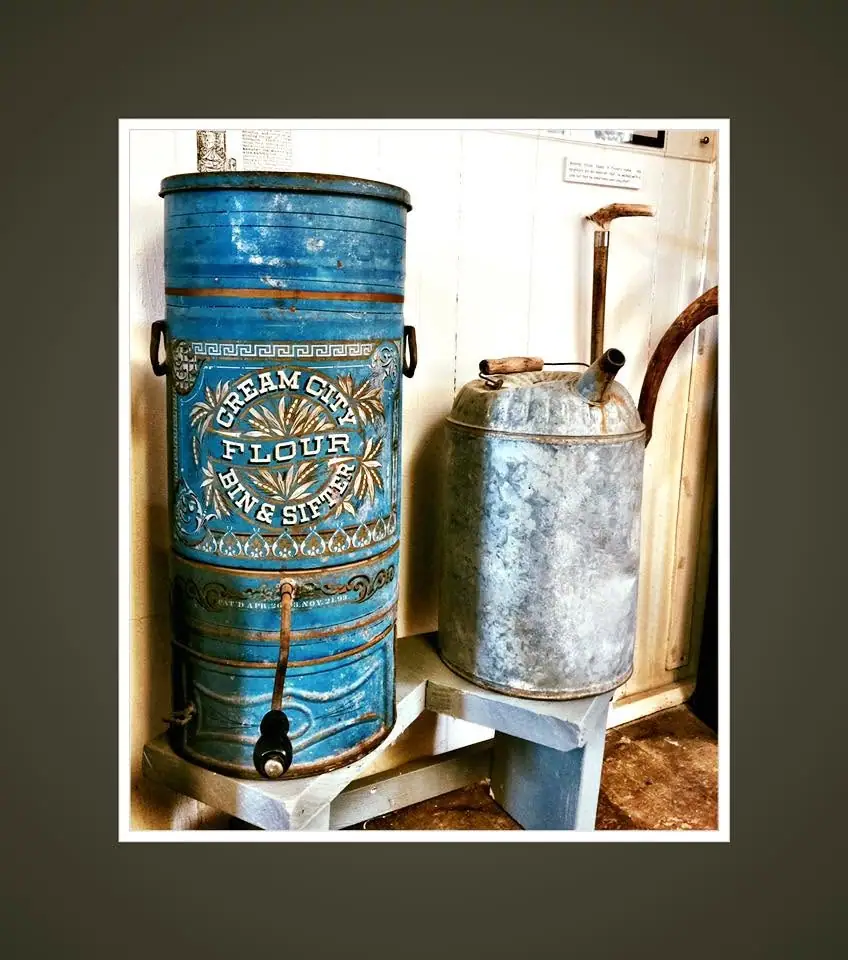 A blue flour bin and sifter stands beside a galvanized metal container in a small indoor exhibit.