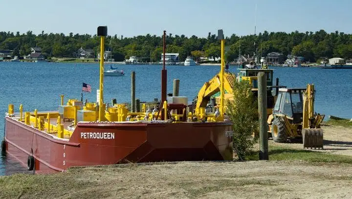 A red work barge with yellow machinery sits beside a shoreline, with a backhoe and houses across the water.
