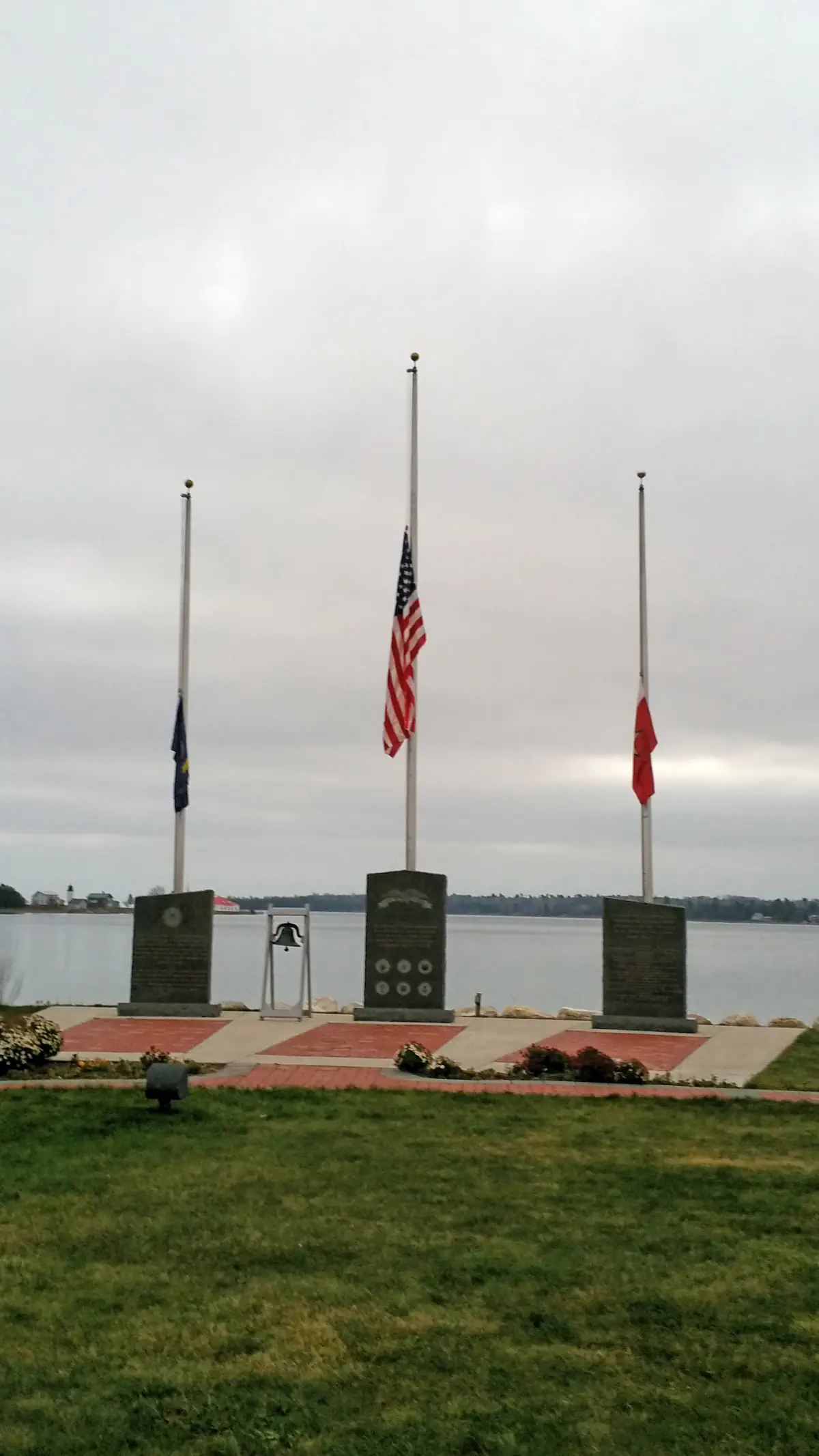 Waterfront memorial with three flagpoles, stone plaques, a bell, and flower beds under an overcast sky.