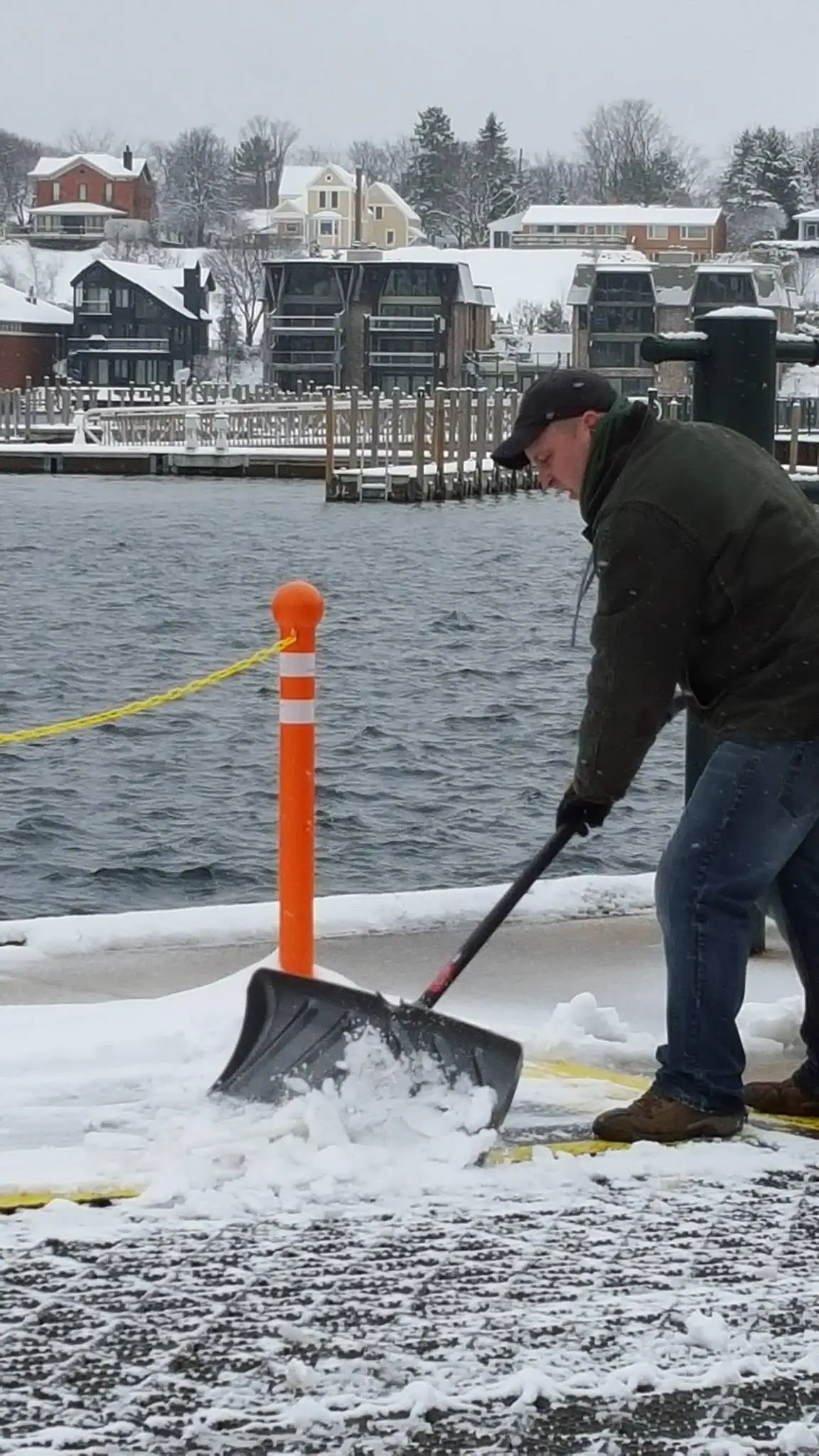 A person shovels snow beside a waterfront, with docks and houses visible across the water.