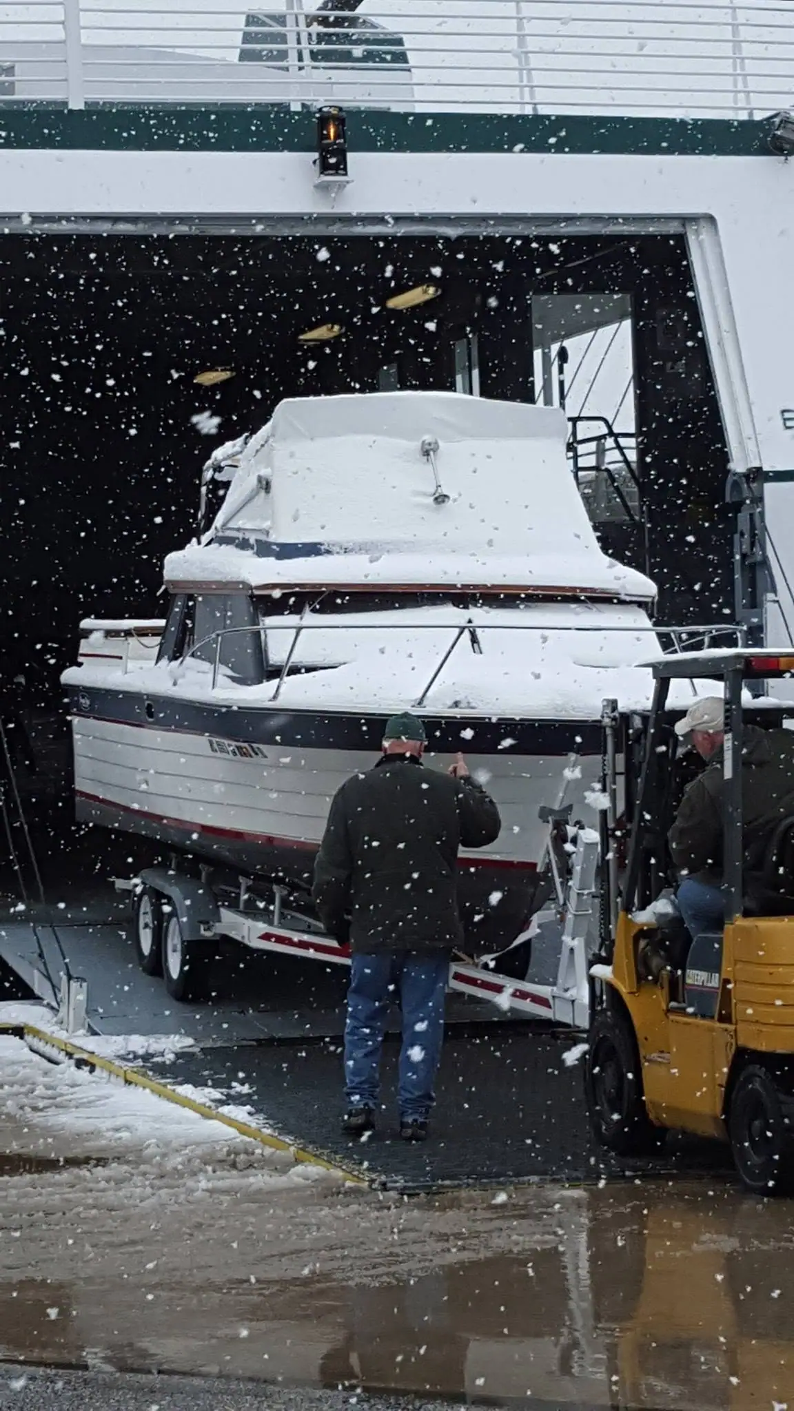A snow-covered cabin boat is loaded on a trailer aboard a large vessel while a forklift and two people stand nearby.