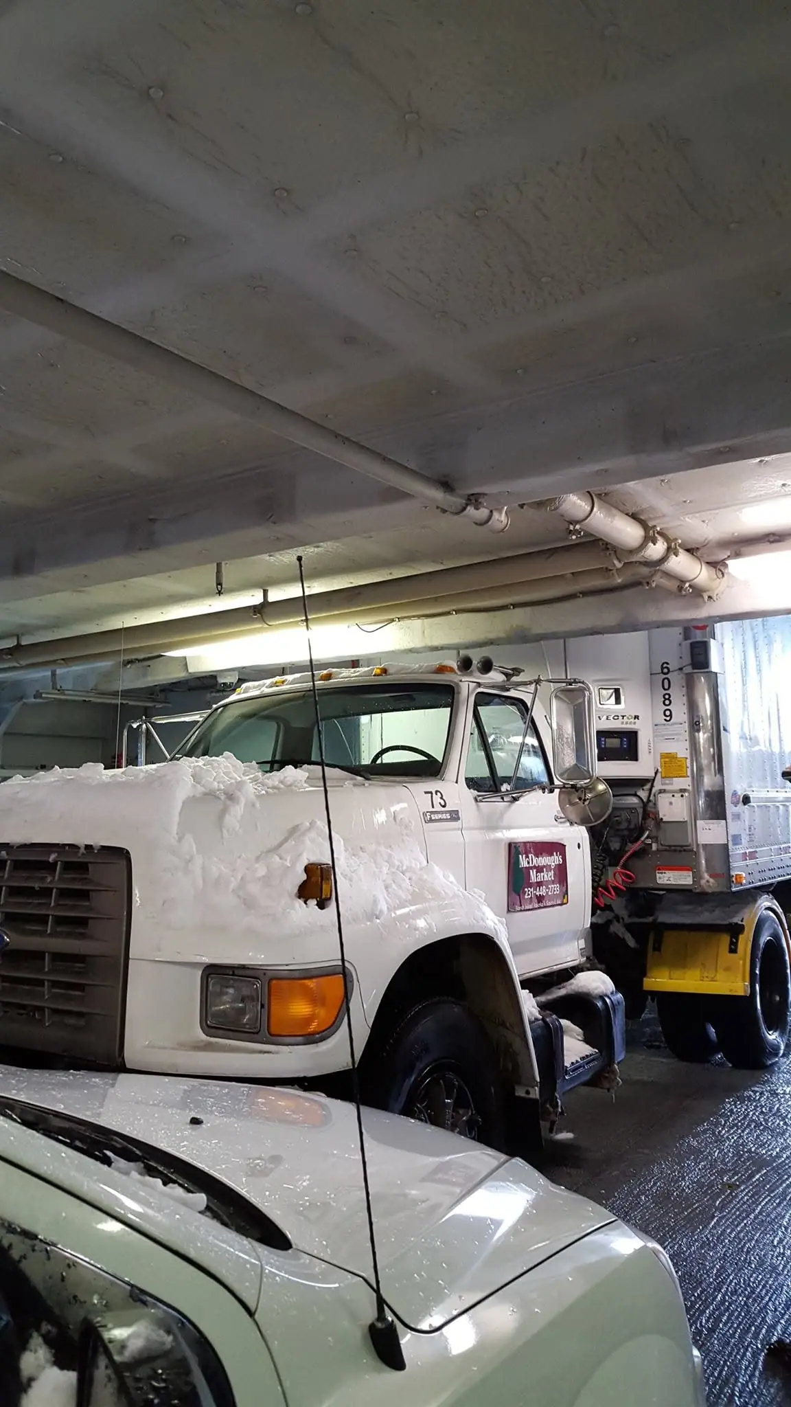 A snow-covered white box truck is parked indoors beside a trailer and another vehicle.