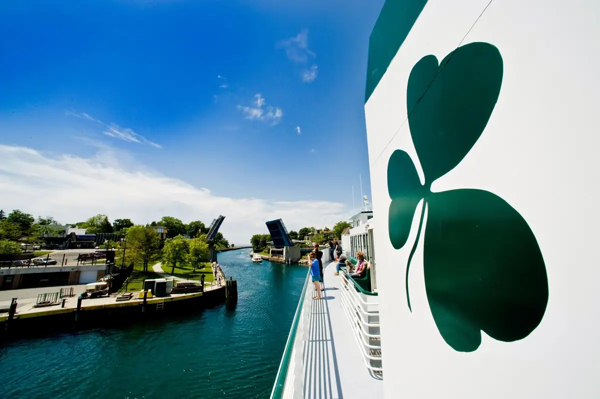 Passengers stand on a ferry deck beside a large green shamrock emblem as raised bridges span a narrow waterway.