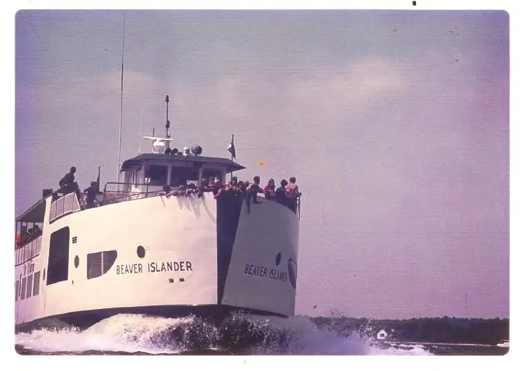 A passenger ferry with people on deck cuts through the water near a wooded shoreline.