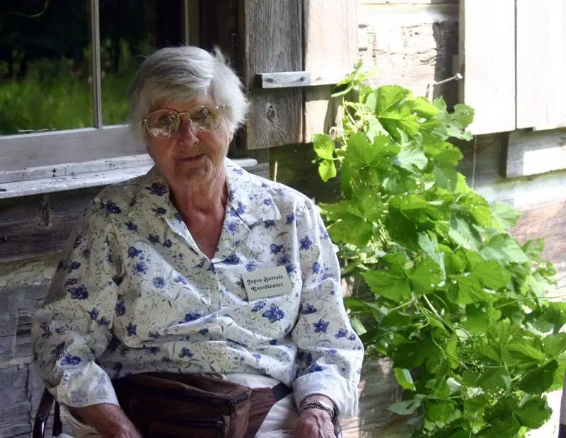 An older woman wearing glasses and a floral blouse sits outdoors beside a leafy vine and weathered wooden building.