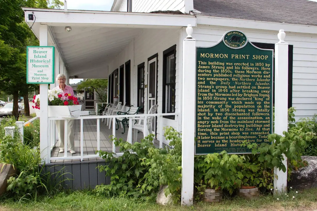 A woman holds a flower-filled planter on the porch of a white wooden museum and print shop.