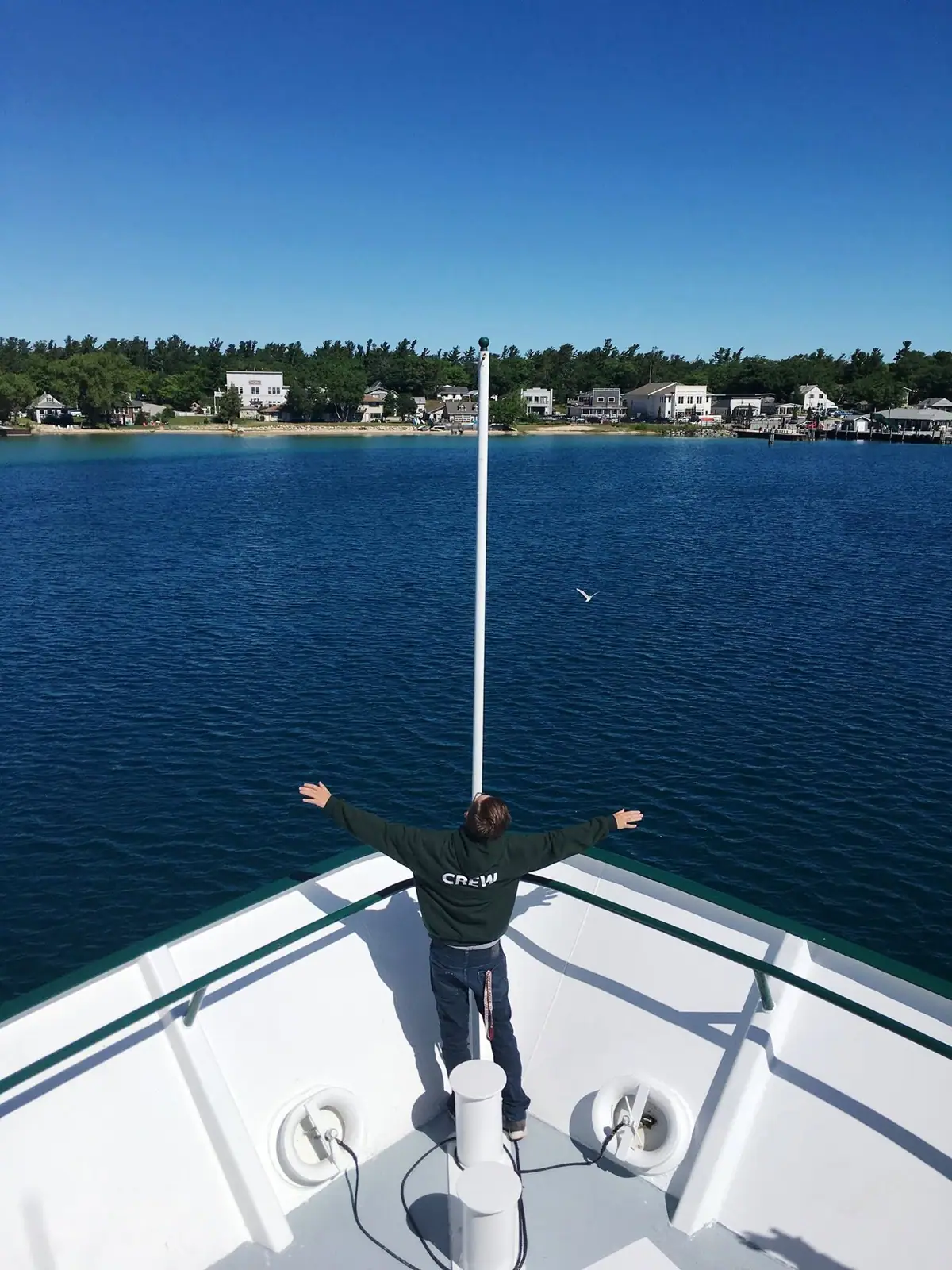 Crew member stands with arms outstretched on a vessel’s bow, facing a tree-lined shoreline and waterfront buildings.