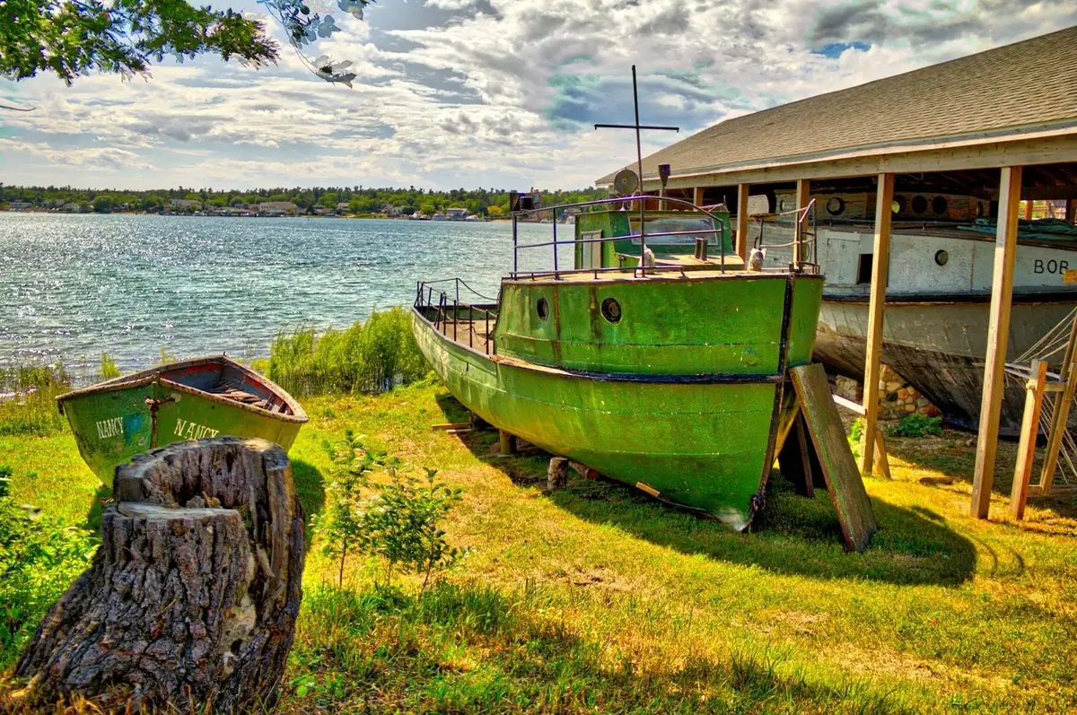 Two green boats rest on shore beside a covered boat shelter, with sparkling water and a distant tree-lined shoreline beyond.