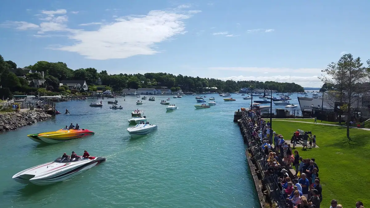 A crowded waterfront overlooks numerous speedboats moving through a channel, with wooded shorelines and houses beyond.