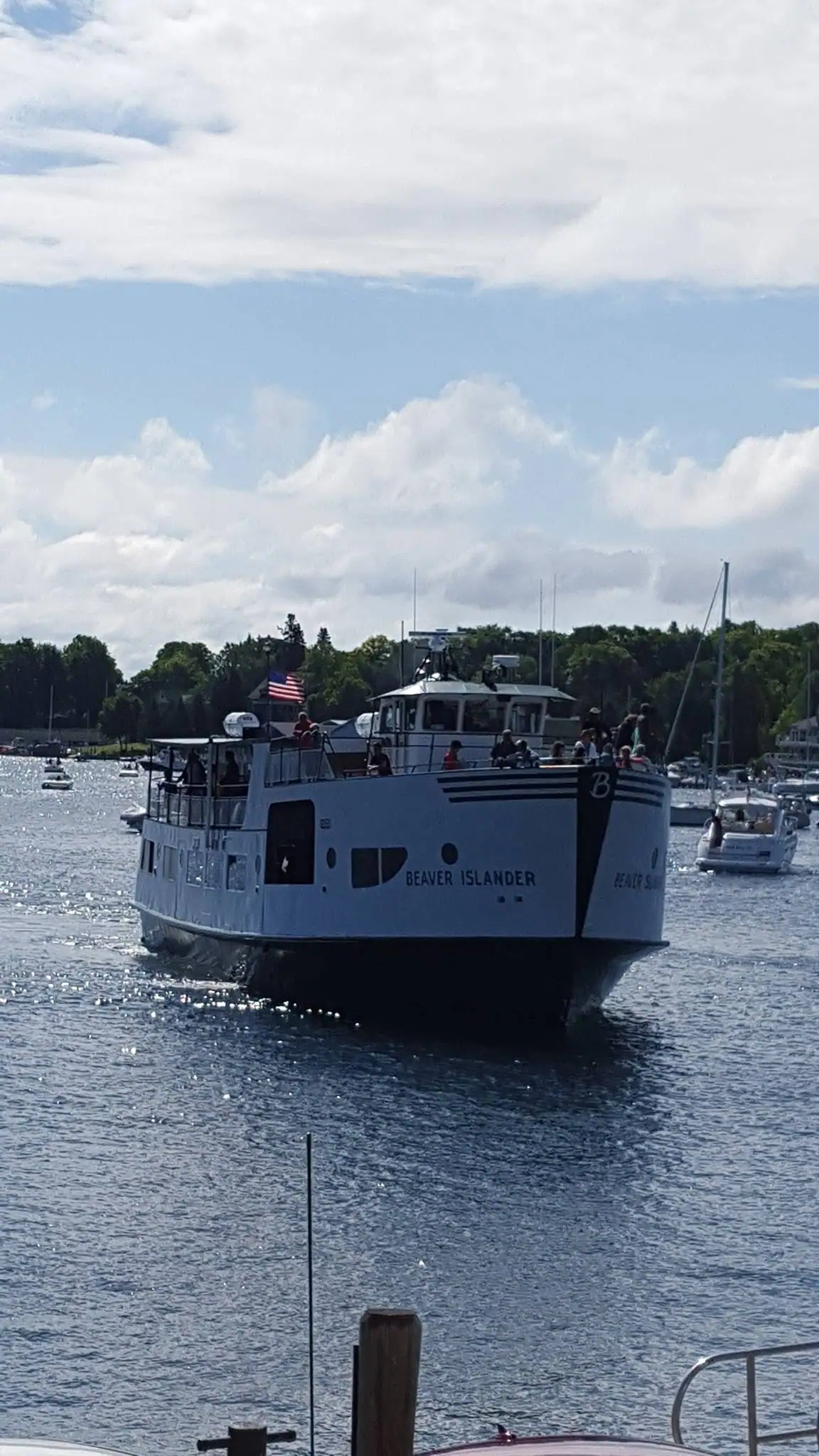 A passenger ferry approaches through sunlit water, with people on deck and smaller boats near a wooded shoreline.