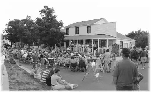 A large outdoor crowd gathers near a porch while people sit in lawn chairs and on the ground.