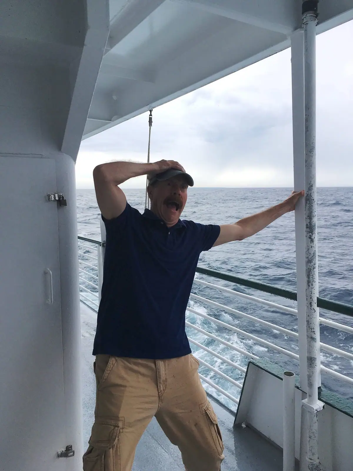 A mustached man in a baseball cap poses on a passenger vessel's deck above choppy open water.