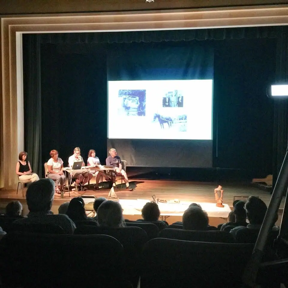 A panel of five people sits on a theater stage before a projected slide while an audience watches.