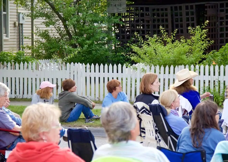 People seated in lawn chairs outdoors beside a white picket fence and a house.