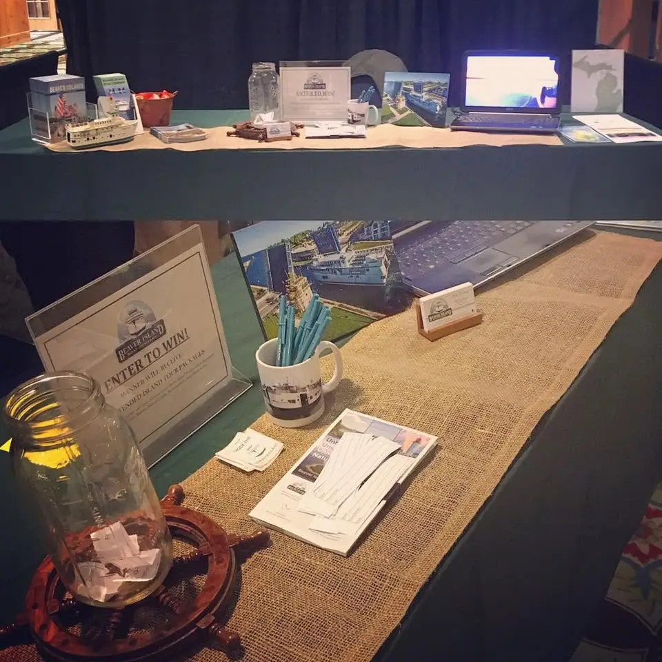 Promotional booth display with a model passenger vessel, raffle jar, brochures, laptop, and printed materials on a table.