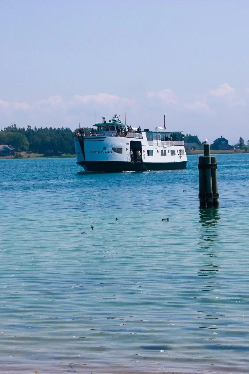 A white passenger ferry passes a wooded shoreline, with dock pilings in the foreground.
