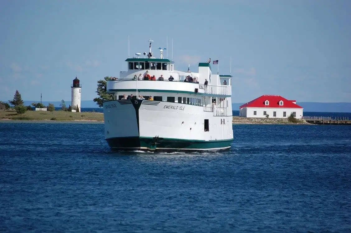 A white-and-green ferry passes a lighthouse and a red-roofed waterfront building.