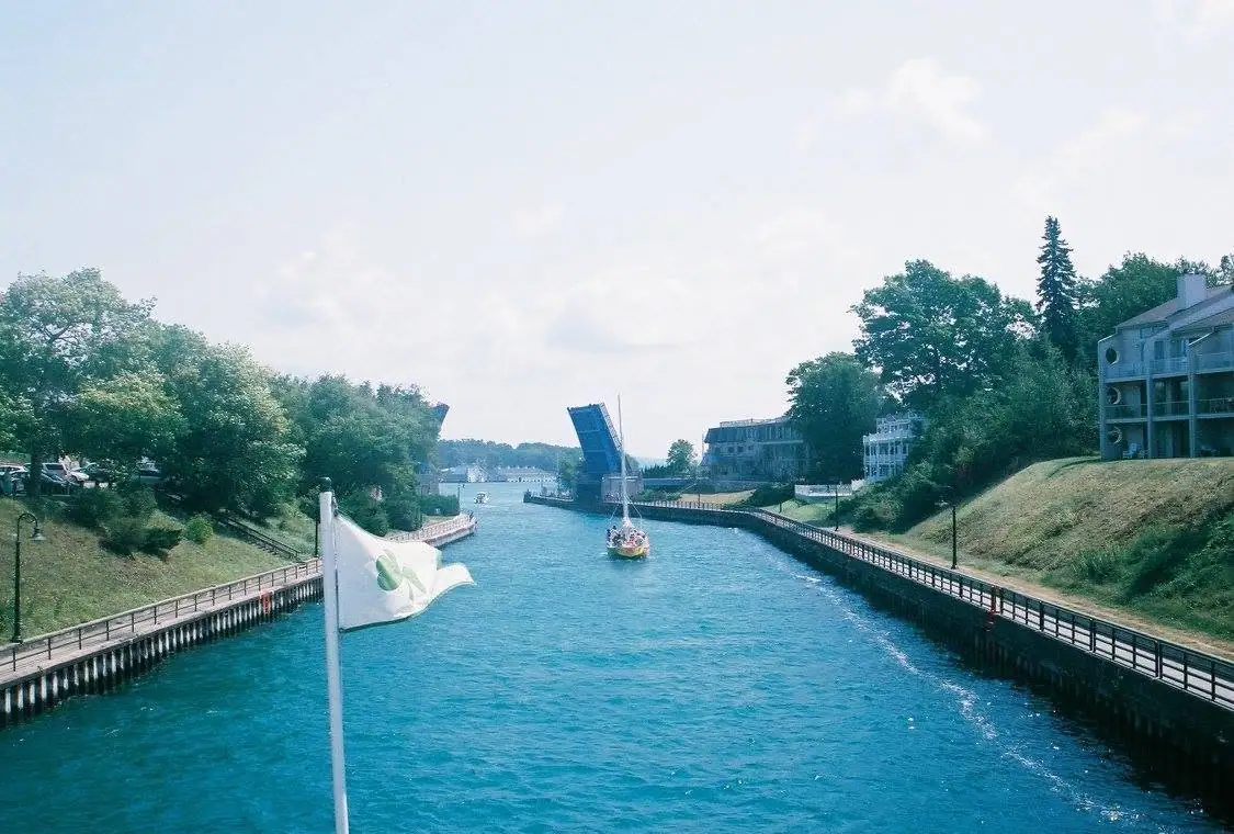A raised drawbridge spans a channel beside a sailboat, waterfront walkways, trees, and a white flag with a green emblem.