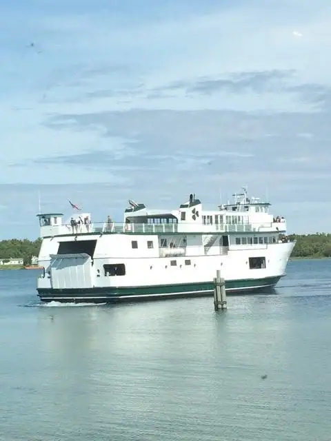 A white passenger ferry with a dark green stripe moves across calm water near a wooded shoreline.