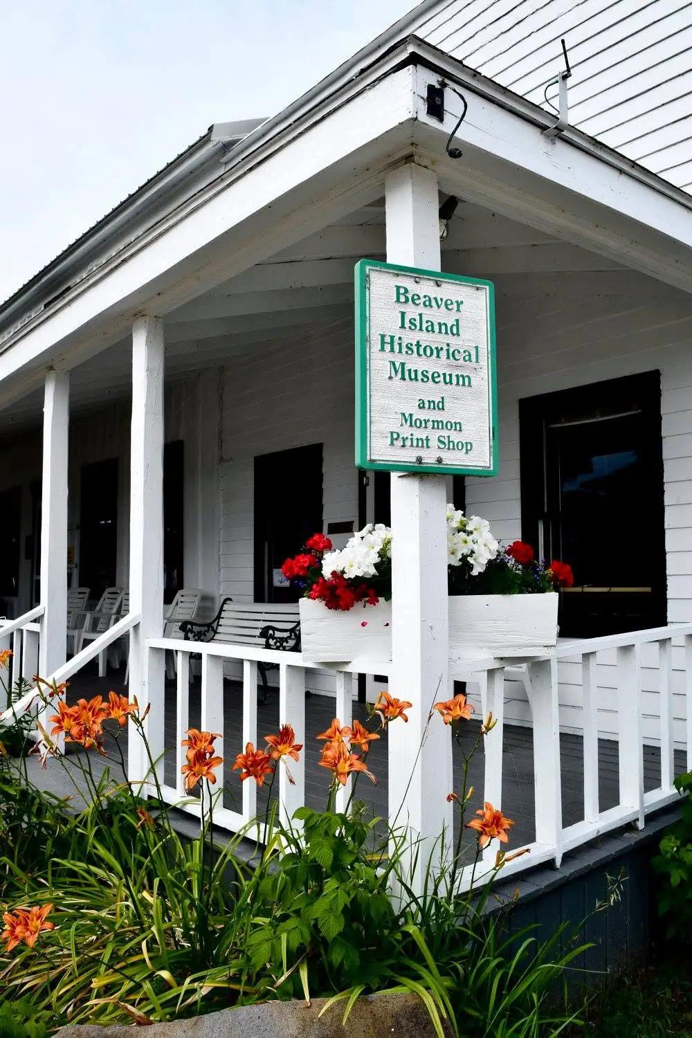 White wooden building with a covered porch, flower boxes, orange lilies, and a green-bordered sign.