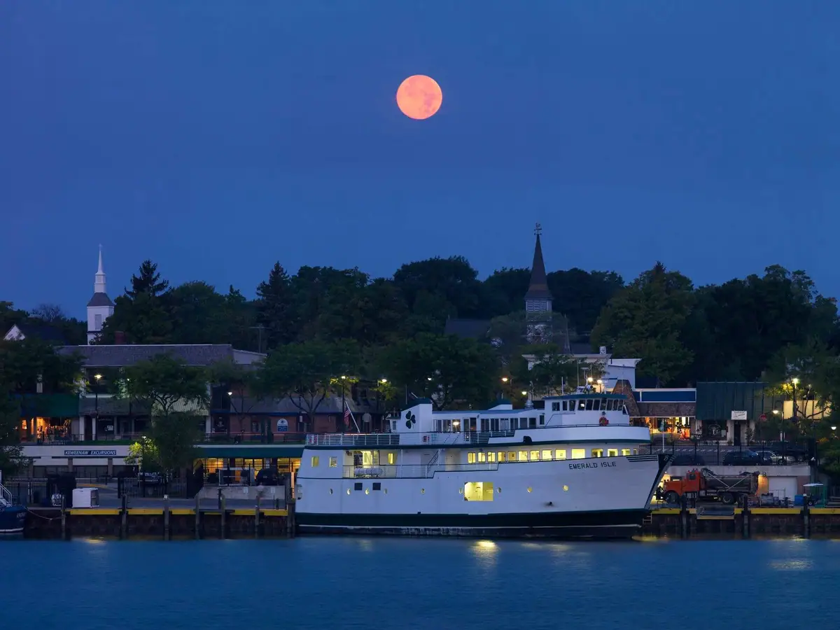 A lit ferry is docked beneath a reddish full moon, with church steeples and tree-covered shoreline behind it.