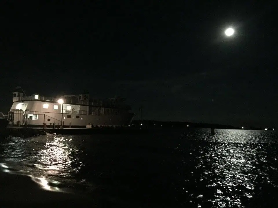 A lit ferry sits beside a pier at night as moonlight shimmers across the water.