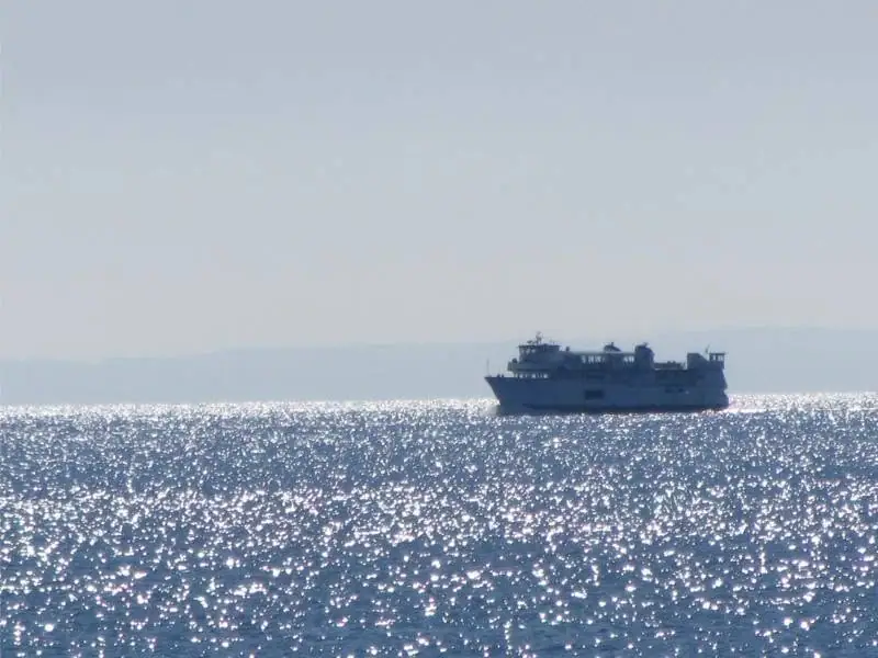 A large ferry travels across intensely sparkling open water beneath a pale, hazy sky.