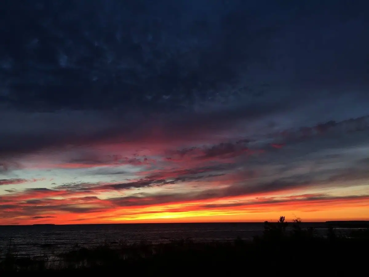A vivid orange sunset glows beneath dark clouds over rippling water and a silhouetted shoreline.