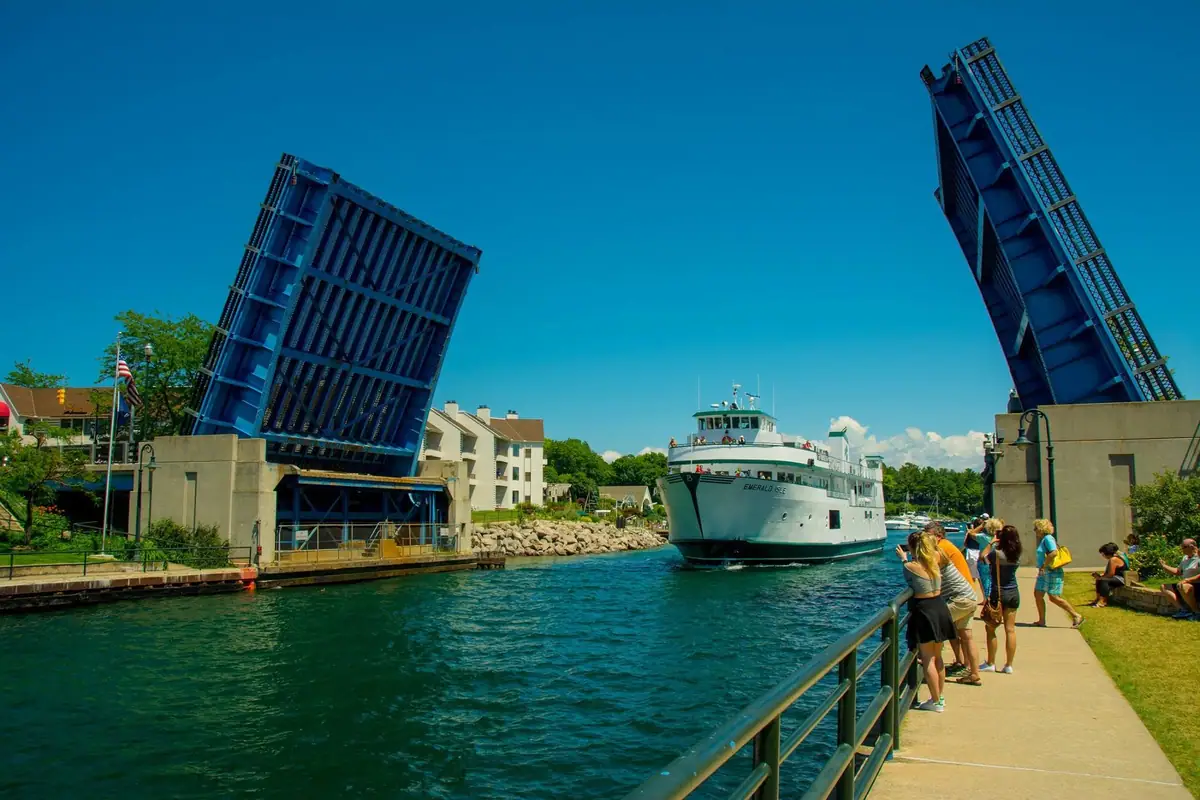 A white ferry passes through a raised blue drawbridge as spectators watch from the walkway.