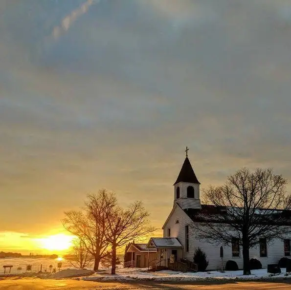 White church with a cross-topped steeple beside a snowy road at sunrise.