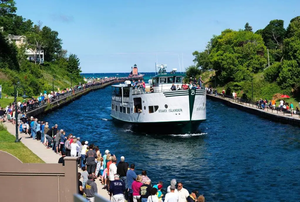 A passenger ferry moves through a channel lined with walkways and crowds, with a lighthouse and open water beyond.