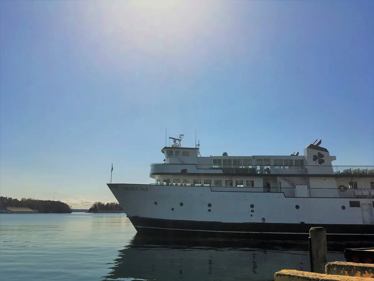 Large white passenger ferry beside a pier, with calm water, wooded shoreline, and a bright blue sky.