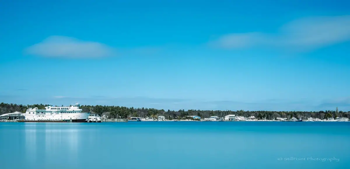 A white ferry rests at a waterfront dock across calm blue water, with homes and trees along the shore.