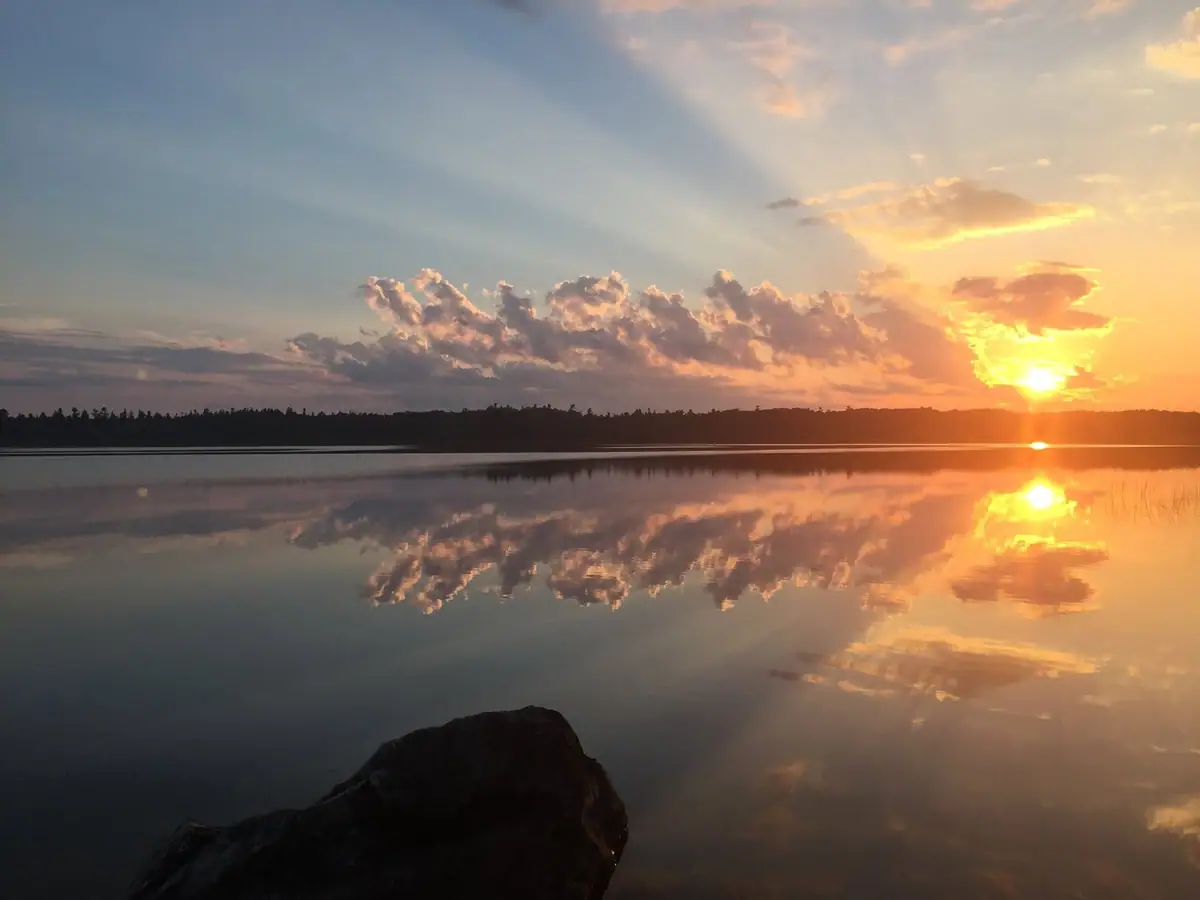 Sunset glows over calm water, reflecting clouds and a wooded shoreline beyond a dark foreground rock.
