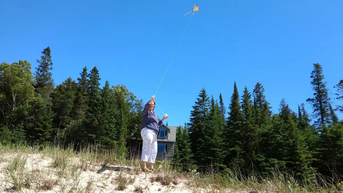 A woman flies a kite from a grassy sandy dune beside a house and dense forest.