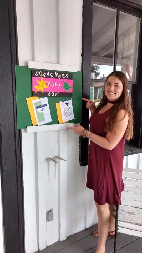 A woman presents a green sign advertising a scavenger hunt outside a building.