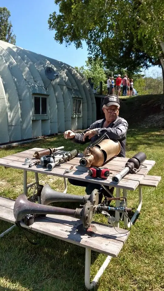 A man works on large metal horns beside picnic tables while people gather near a curved building.