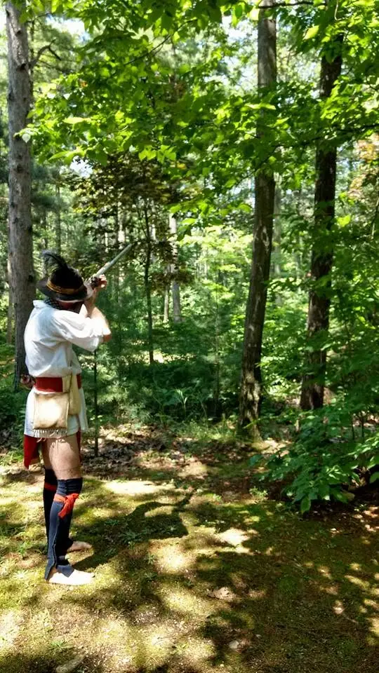 A person in historical-style clothing aims a long rifle in a sunlit forest.